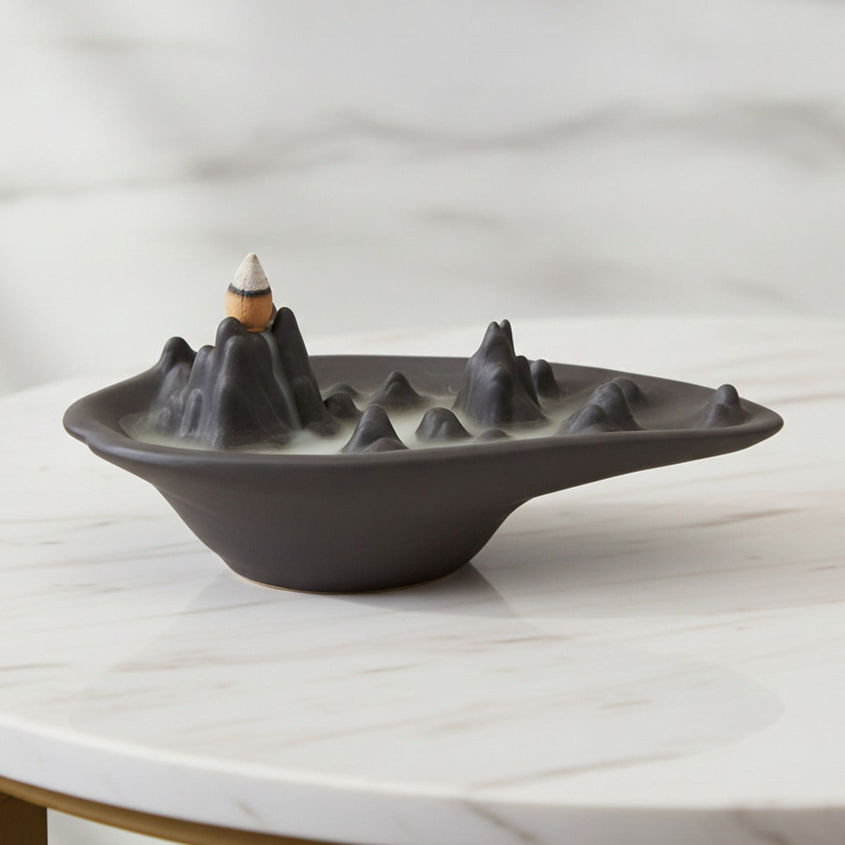 Mountain scape backflow incense burner in matt charcoal ceramic with a lit cone and smoke pooling around the peaks, on a white marble table