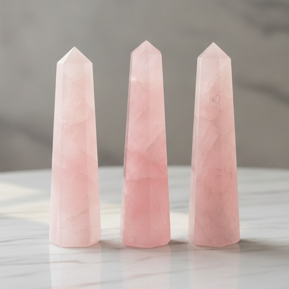 Three polished rose quartz obelisk points in soft pink, standing on a white marble table