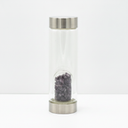 Amethyst crystal glass water bottle with crystal chips inside on a clean white surface against a white wall