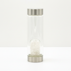 Clear quartz crystal glass water bottle with crystal chips inside on a clean white surface against a white wall