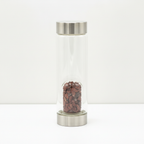 Red jasper crystal glass water bottle with crystal chips inside on a clean white surface against a white wall