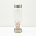 Rose quartz crystal glass water bottle with crystal chips inside on a clean white surface against a white wall
