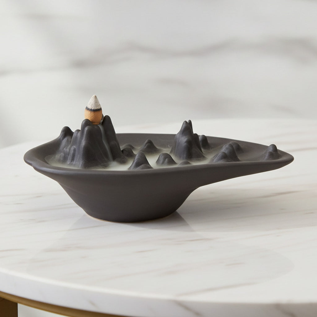 Mountain scape backflow incense burner in matt charcoal ceramic with a lit cone and smoke pooling around the peaks, on a white marble table