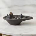 Mountain scape backflow incense burner in matt charcoal ceramic with a lit cone and smoke pooling around the peaks, on a white marble table