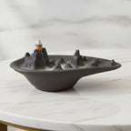 Mountain scape backflow incense burner in matt charcoal ceramic with a lit cone and smoke pooling around the peaks, on a white marble table