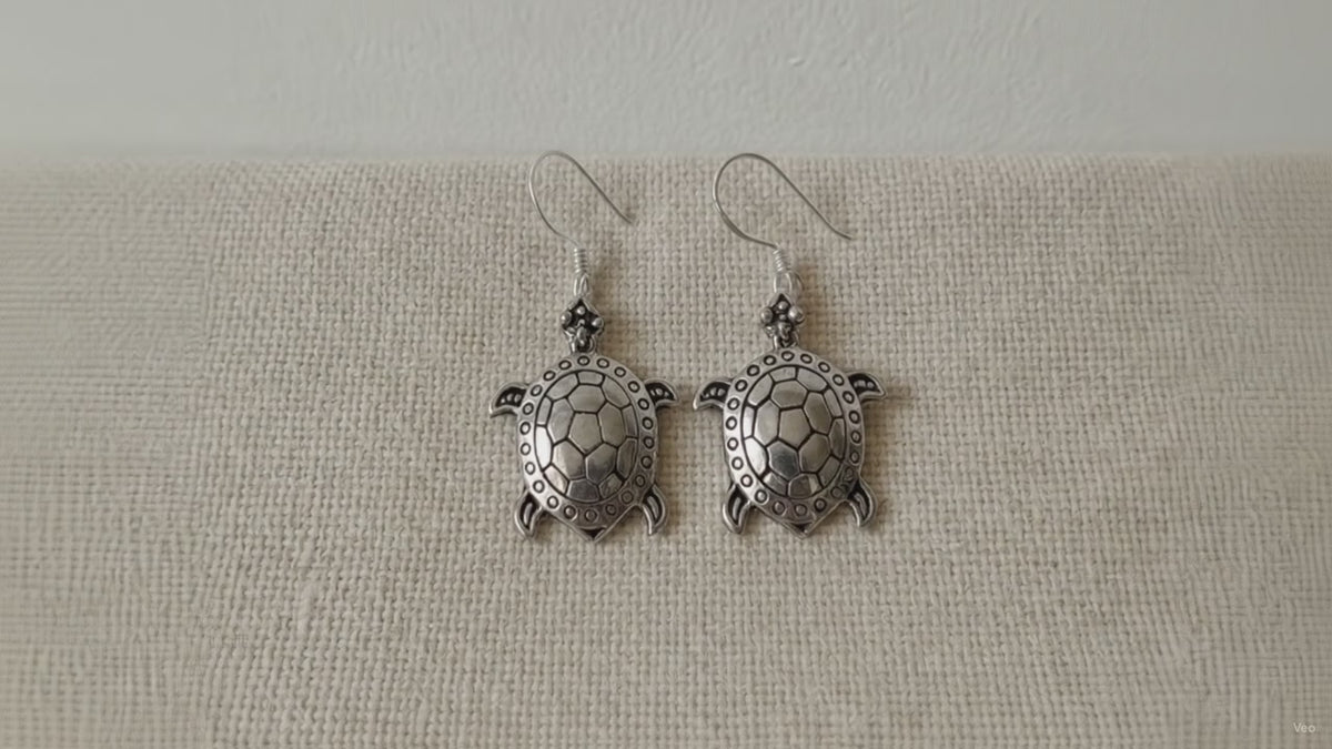 A pair of sterling silver turtle drop earrings on cotton canvas video