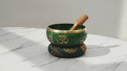 Large green singing bowl set with gold Tree of Life symbols, cushion and wooden striker on marble, slow push-in