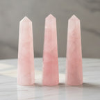 Three polished rose quartz obelisk points in soft pink, standing on a white marble table