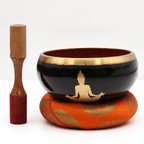 Black singing bowl with a gold seated Buddha on an orange cushion, wooden striker beside it